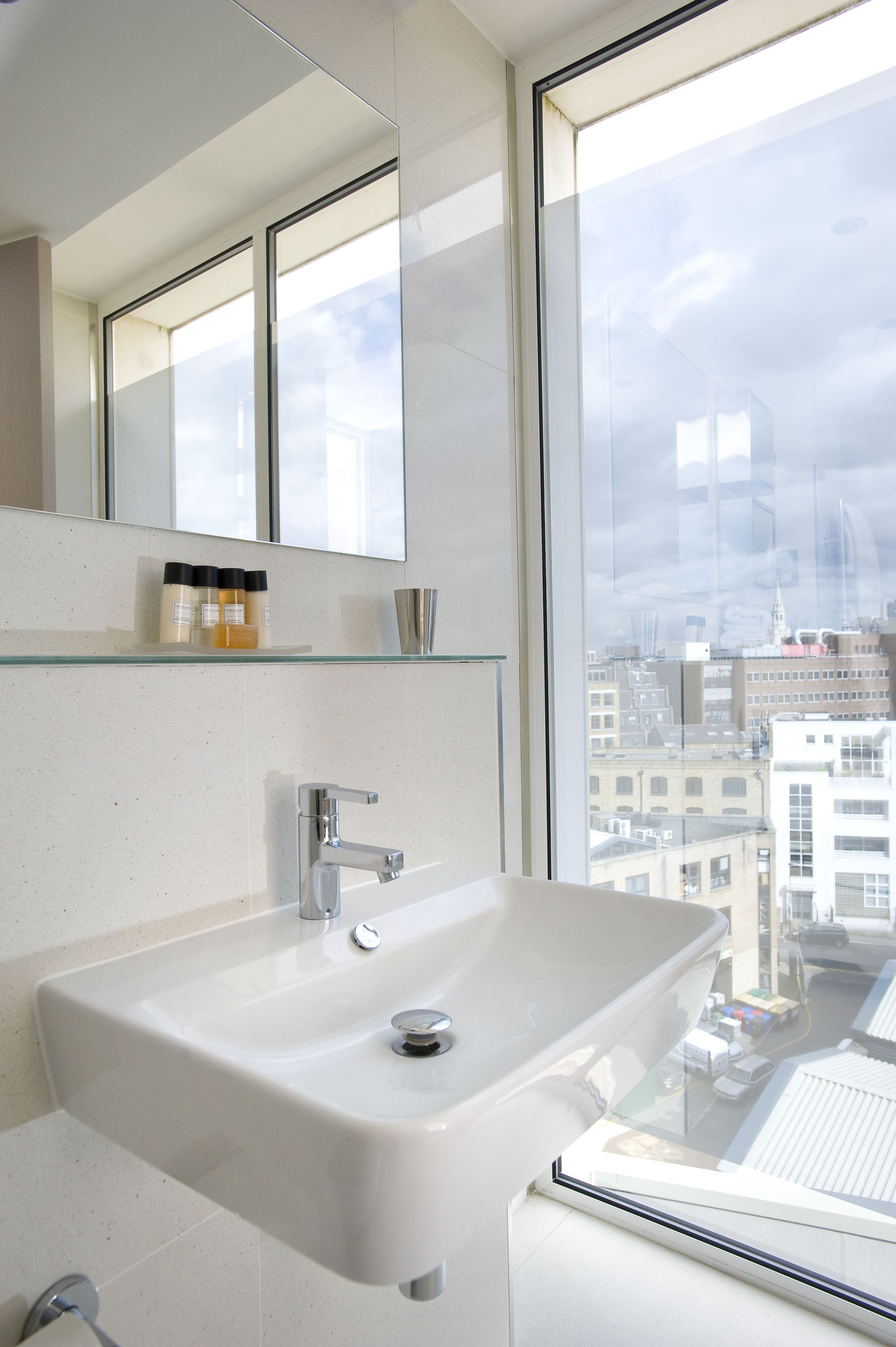A modern bathroom with a large window, a white sink, and a glass shelf with toiletries.