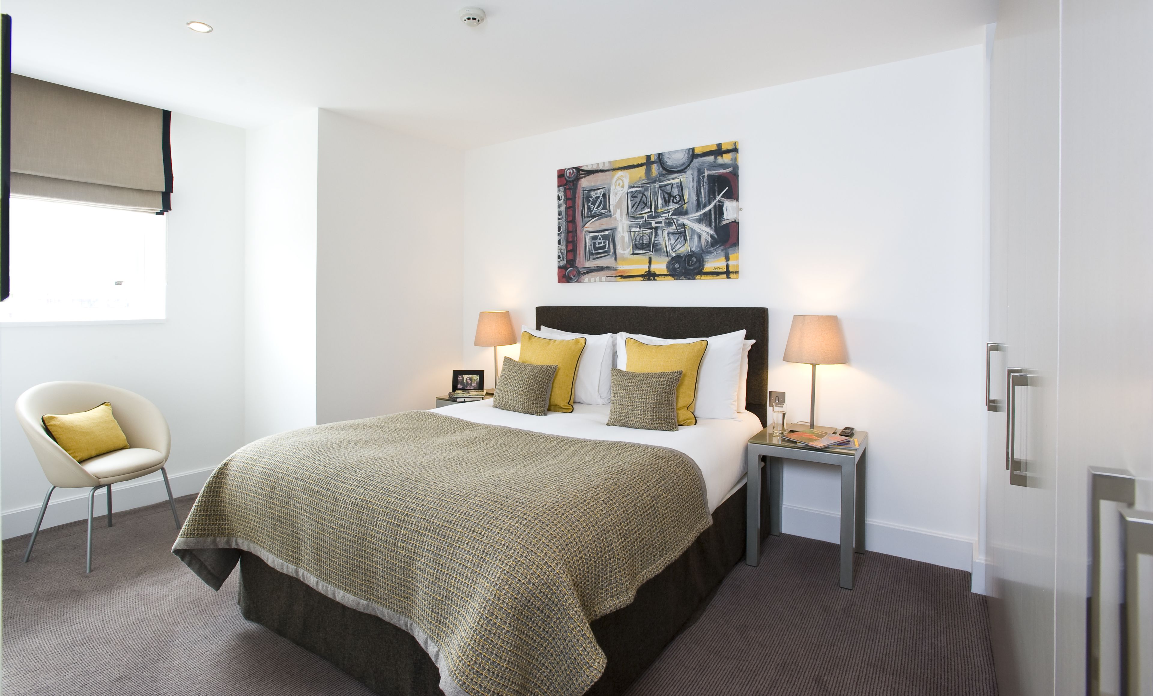 A modern hotel room with a large bed, two bedside tables, a chair, a painting on the wall, and a window with a blind.