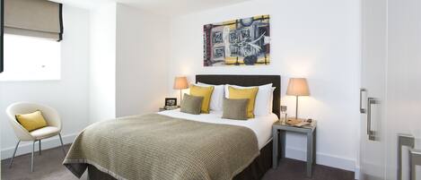 A modern hotel room with a large bed, two bedside tables, a chair, a painting on the wall, and a window with a blind.