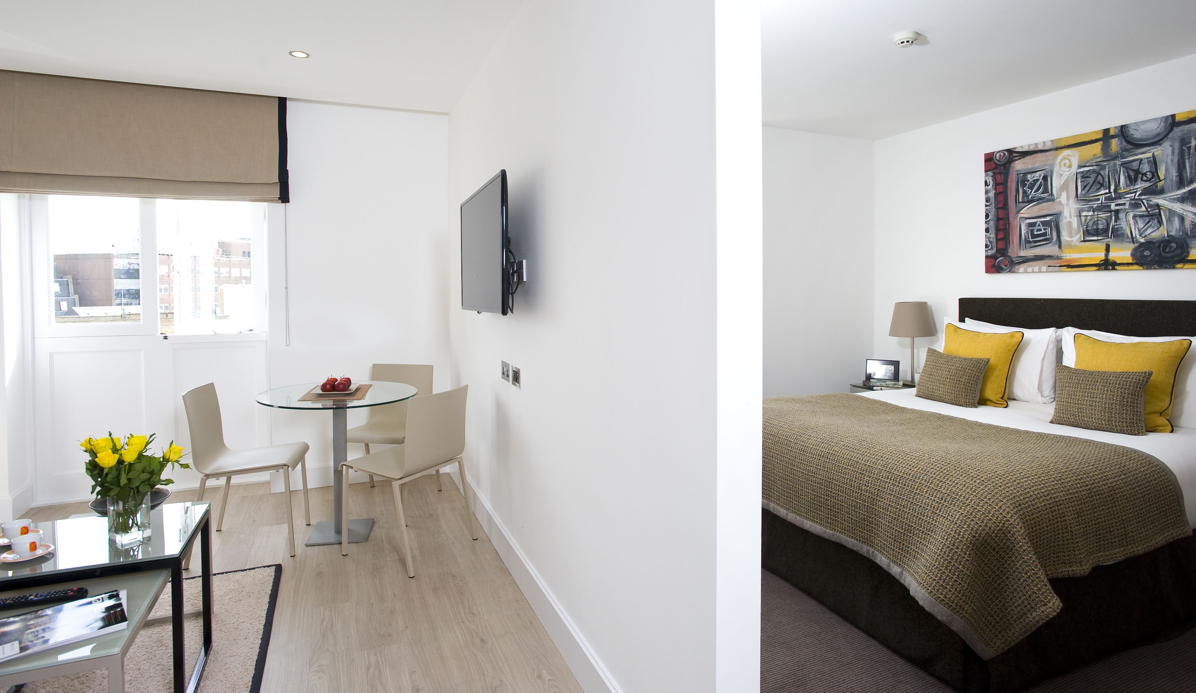 A hotel room with a bed, a TV, a dining area with a table and chairs, and a window with a view of buildings.