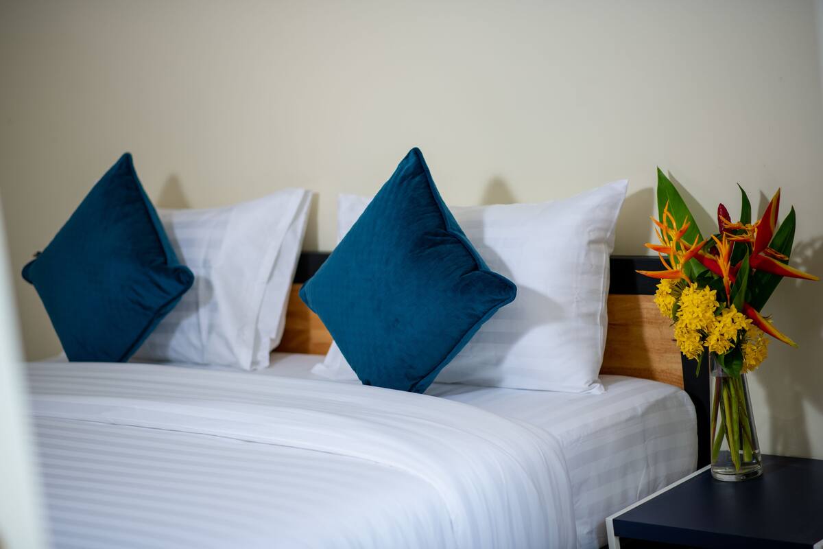 A hotel room with a bed, white bedding, teal pillows, and a vase with yellow flowers on a nightstand.