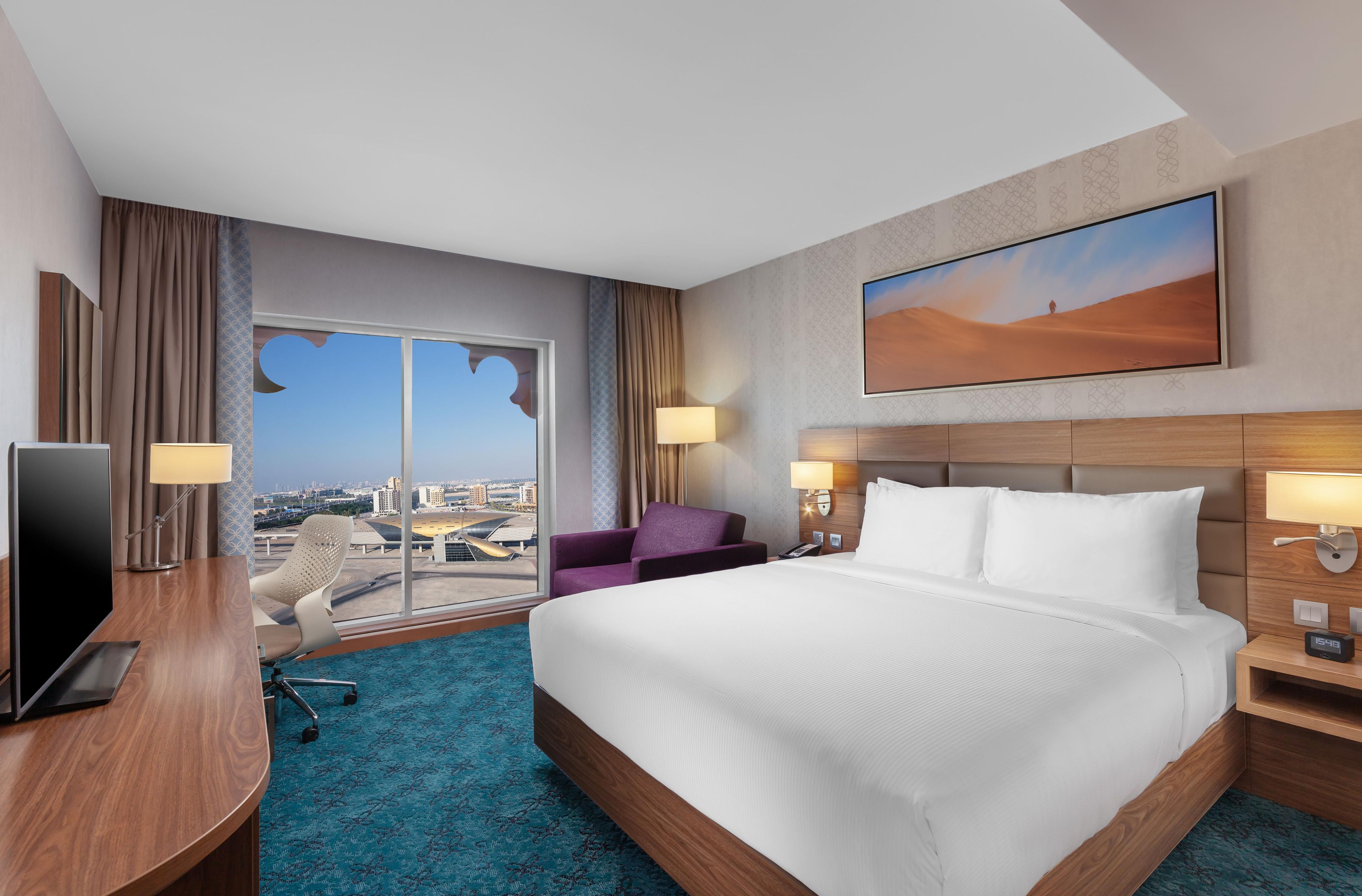 Une chambre d’hôtel avec un grand lit, un bureau avec une télévision, une chaise, un canapé et une vue sur le paysage urbain, visible à travers une grande fenêtre.