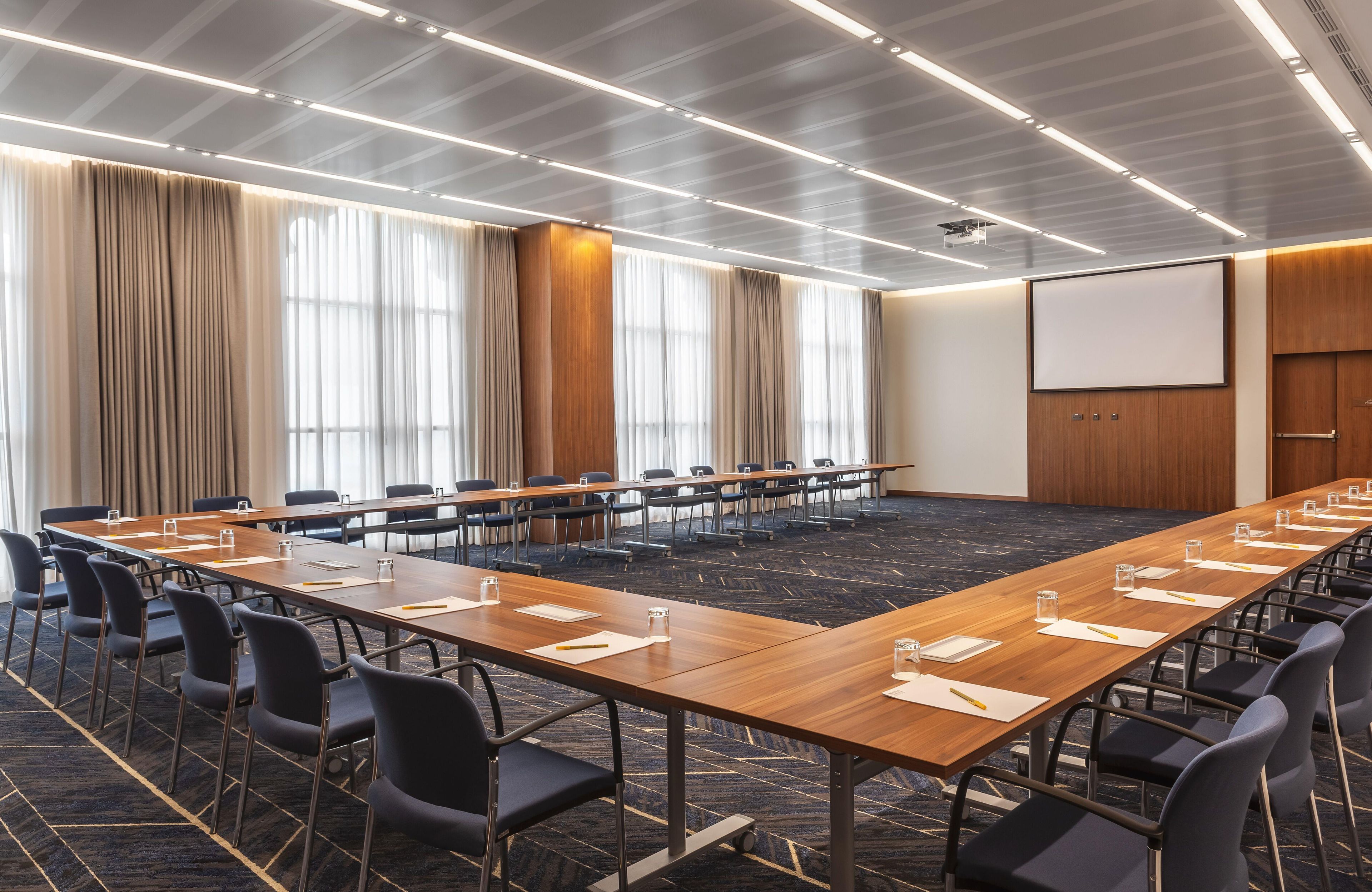 A conference room with a long table, chairs, a projector screen, and a large window with curtains.