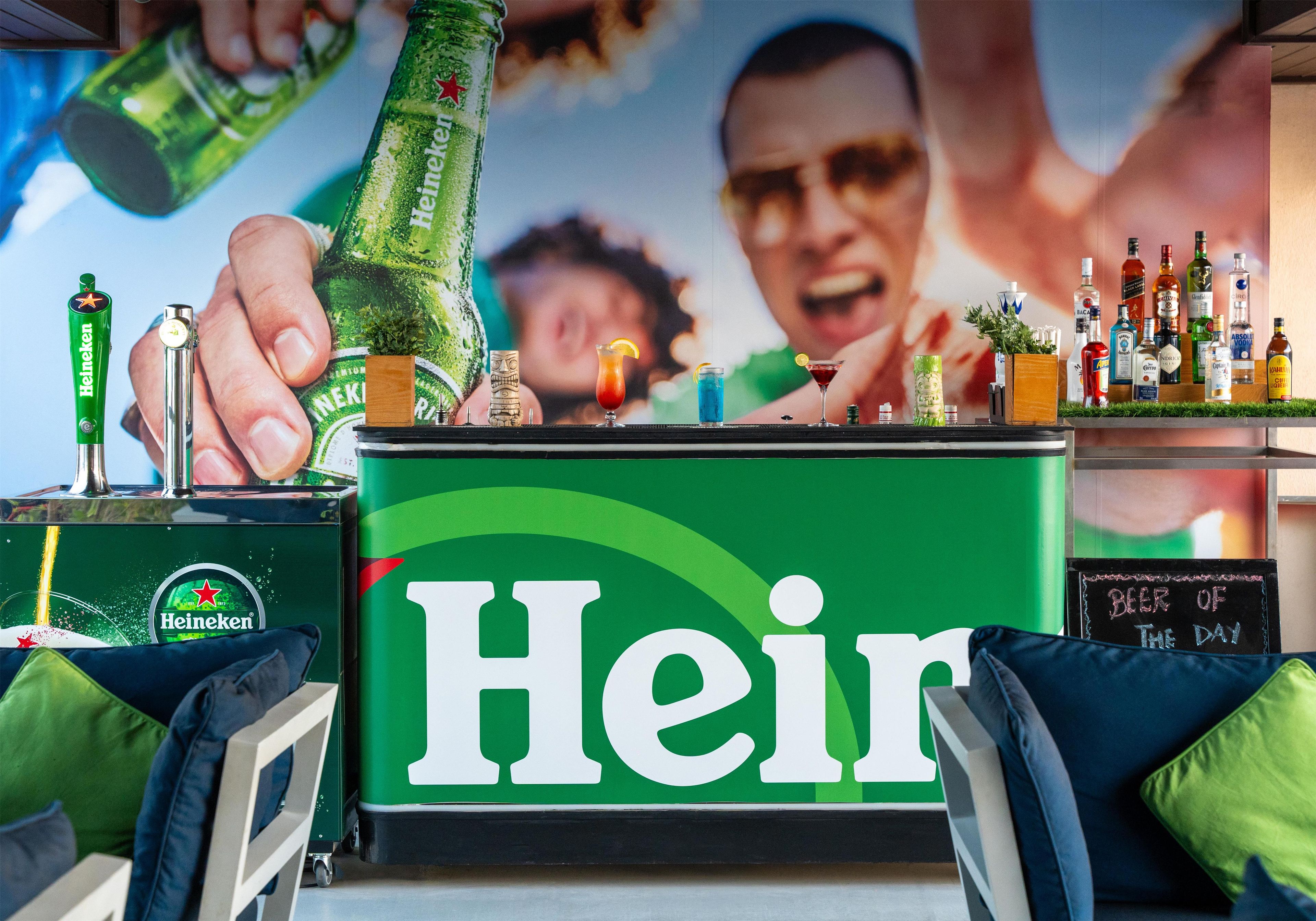 Uma área de bar com um letreiro chamativo da Heineken, variedade de garrafas e um cardápio em lousa.