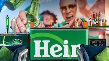 Uma área de bar com um letreiro chamativo da Heineken, variedade de garrafas e um cardápio em lousa.