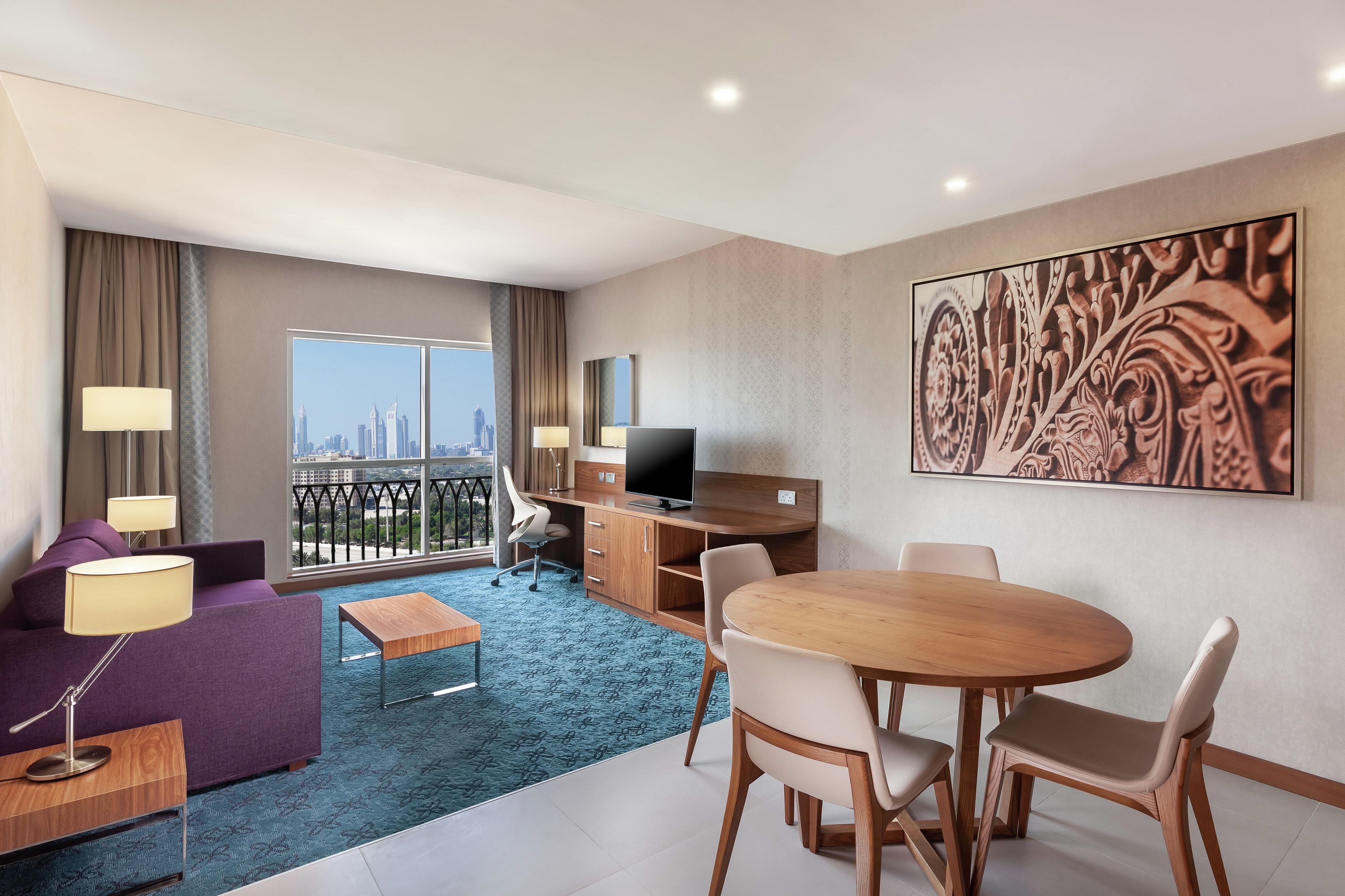 Ein Hotelzimmer mit Blick auf die Stadt, einem Balkon, einem Schreibtisch, einem Fernseher, einem Esstisch und einem großen Wandbild.