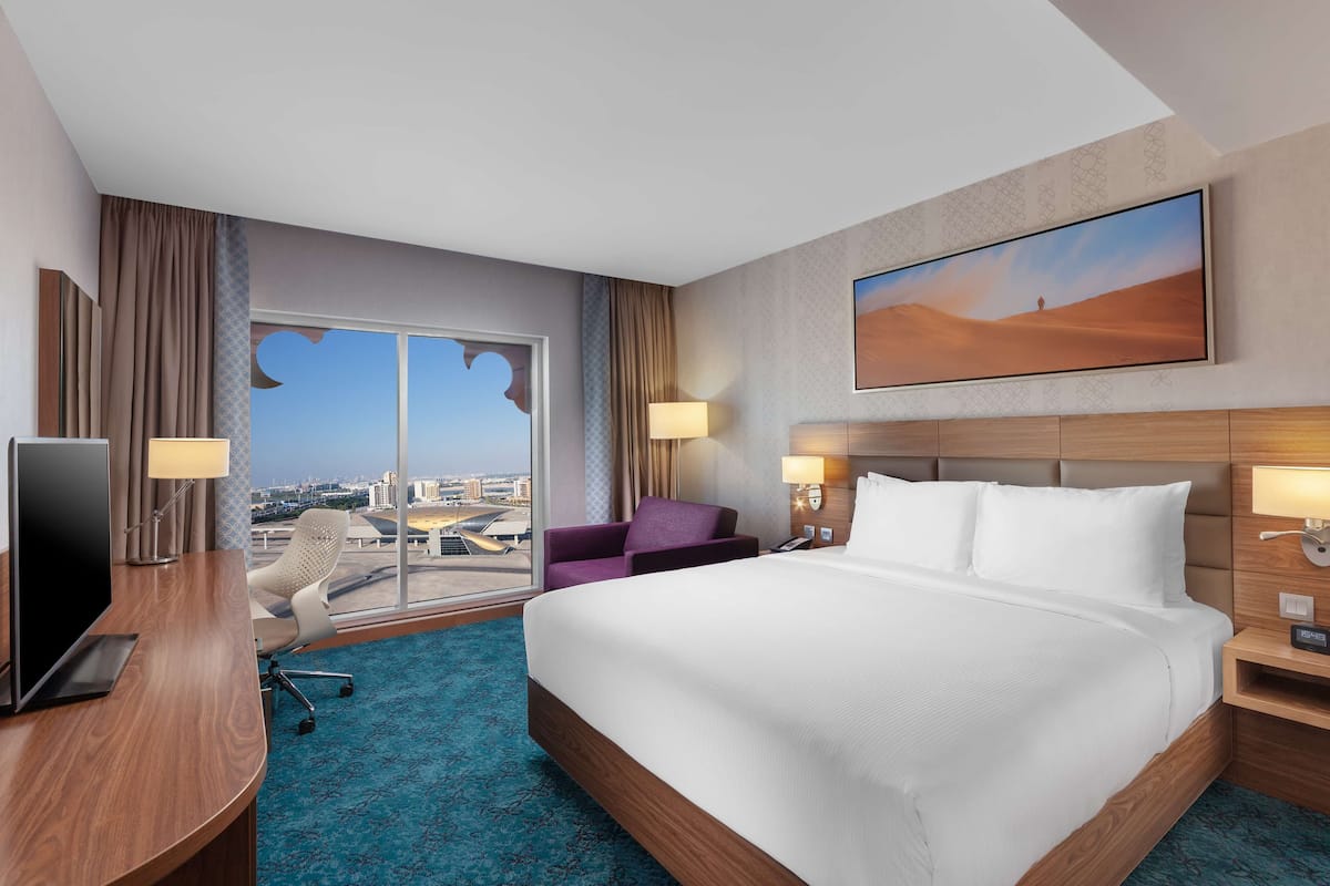 Une chambre d’hôtel avec un grand lit, un bureau équipé d’un ordinateur, une chaise et une vue sur le paysage urbain par une grande fenêtre.