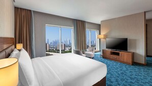 Ein Hotelzimmer mit einem großen Bett, einem Fernseher, einem Schreibtisch und Blick auf eine Stadtlandschaft.
