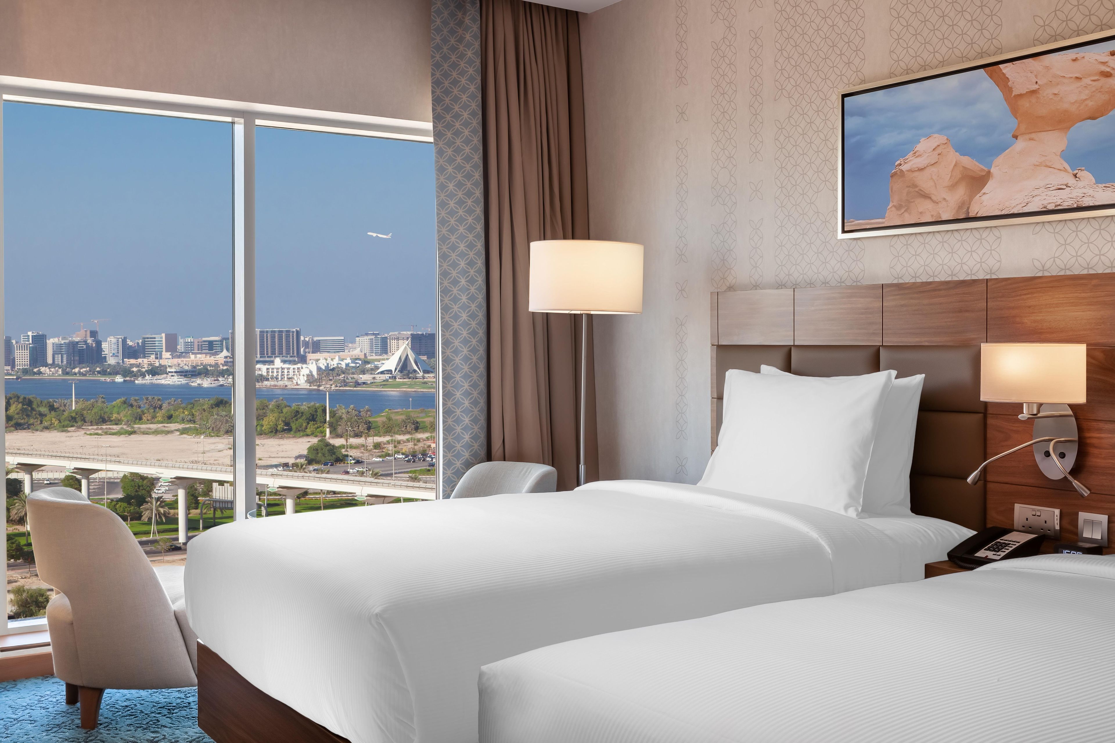 A hotel room with two beds, a city view, a painting, and a desk with a lamp.