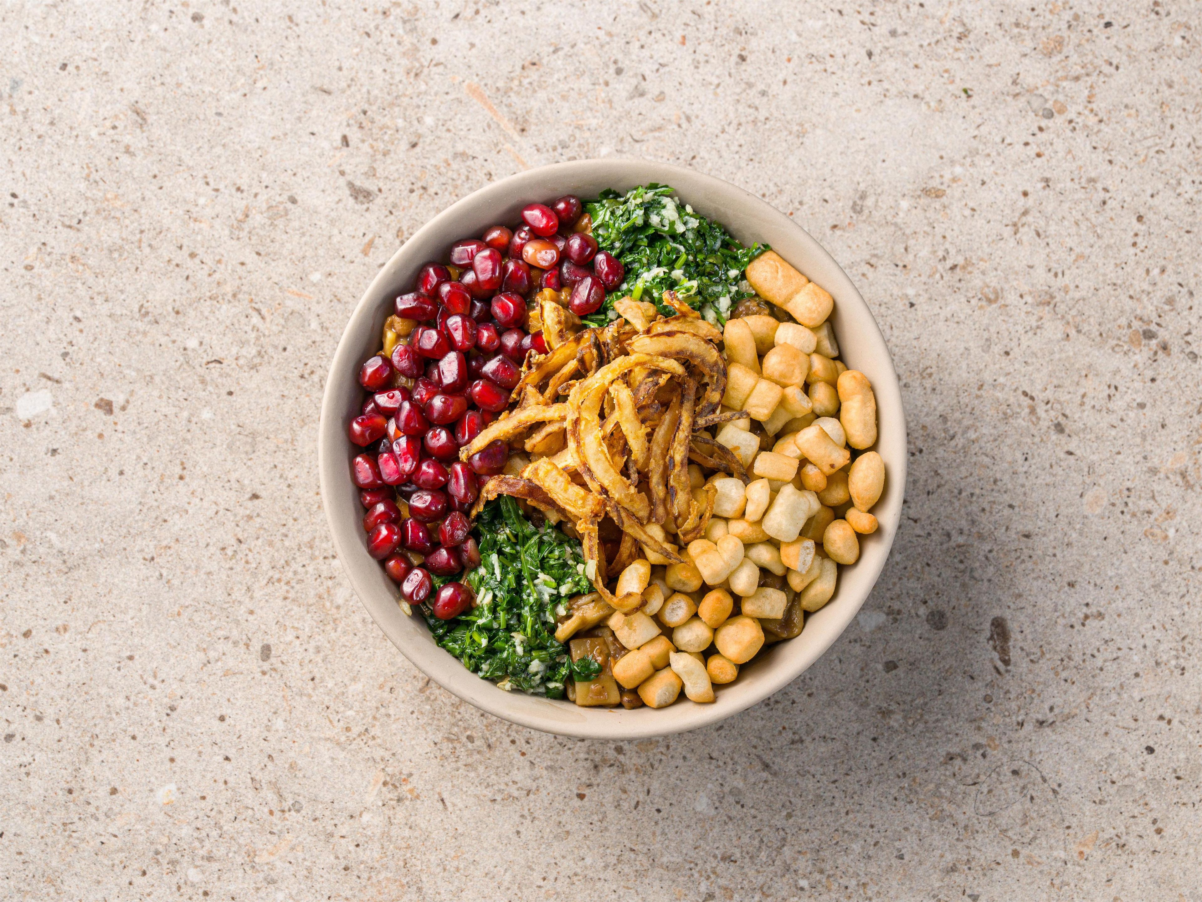 A bowl of food with pomegranate seeds, fried onions, and croutons on a textured surface.
