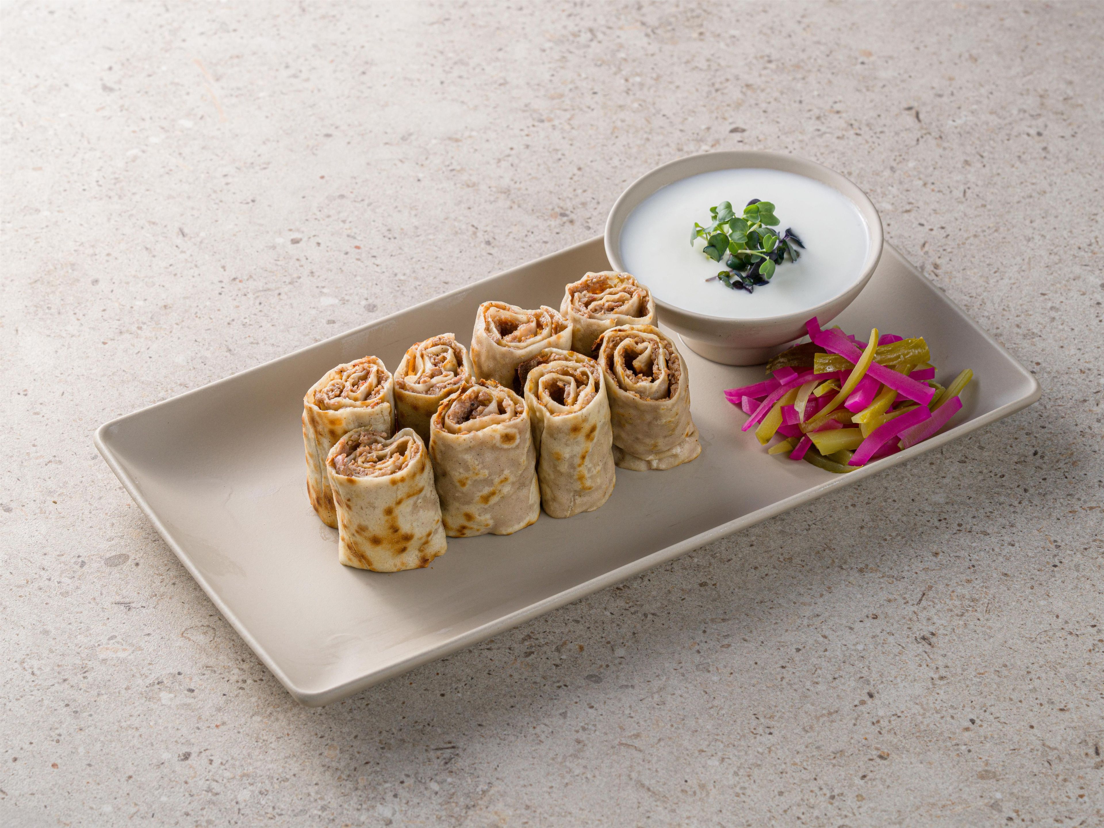A plate with rolled food items, a bowl of dip, and a side of pickled vegetables.