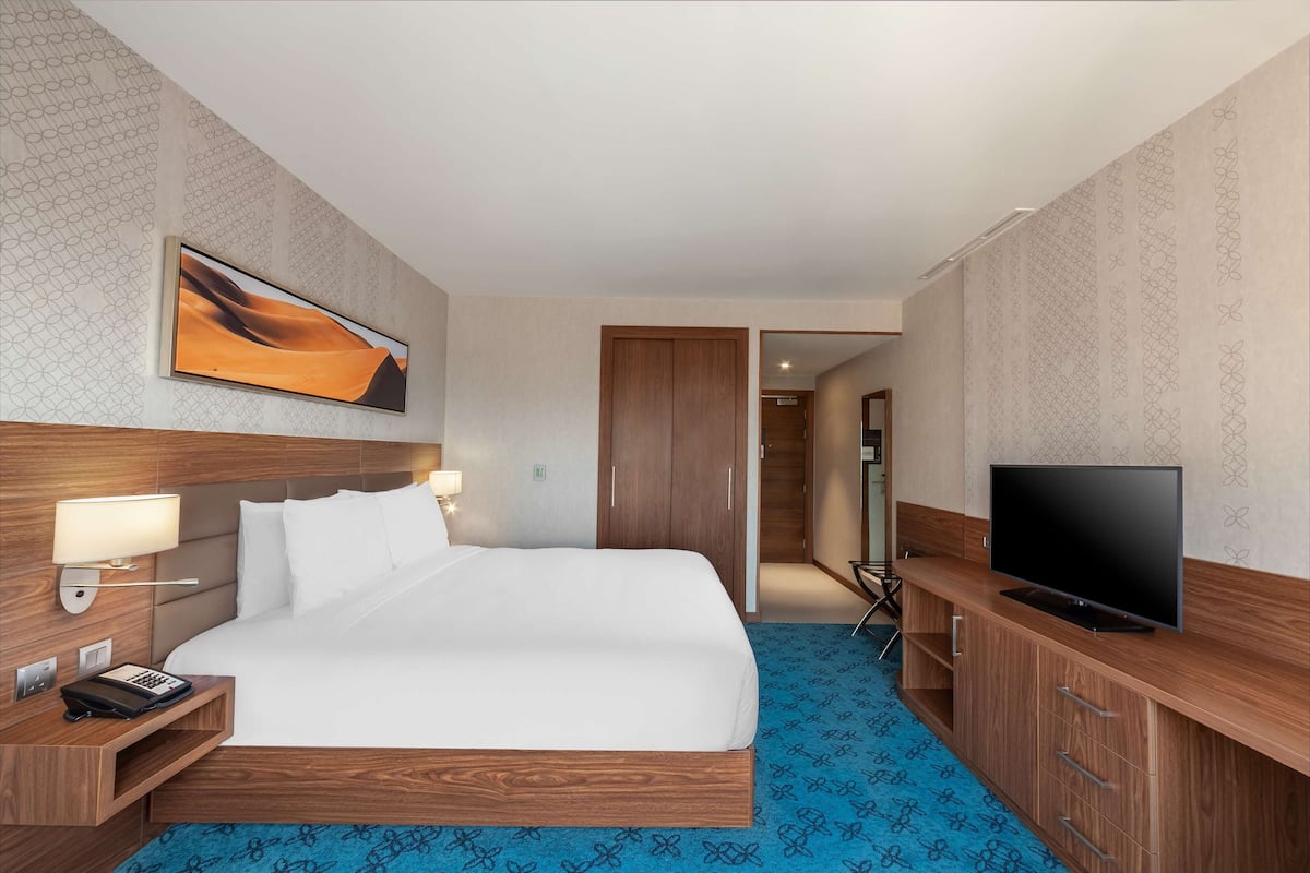 Une chambre d’hôtel avec un grand lit, une tête de lit en bois, un téléviseur à écran plat, un bureau avec une lampe et une moquette à motifs.