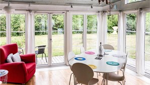Ein heller Raum mit einem roten Sofa, einem runden Tisch mit Stühlen und großen Fenstern.