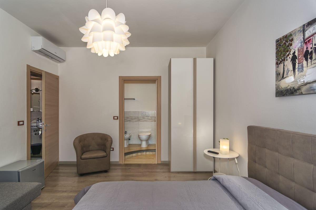 Un dormitorio moderno con cama, sillón, mesita de noche con lámpara, aire acondicionado, armario y baño visible a través de una puerta abierta.
