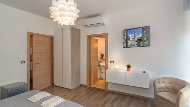 A modern bedroom with a bed, bedside tables, a chair, a pendant light, a radiator, and a painting of a cityscape. - La Candelaria (Pisa)