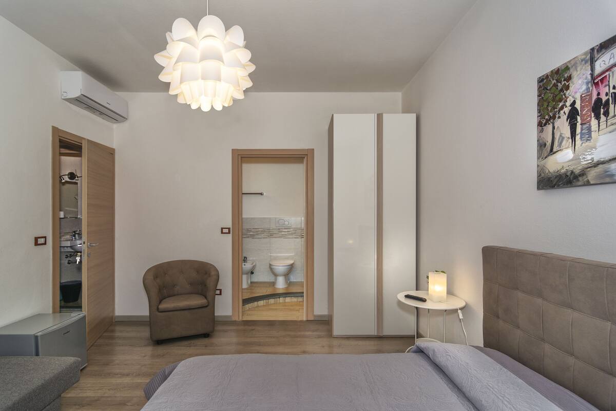 Un dormitorio moderno con cama, una silla, una mesita de noche con lámpara, un armario y un equipo de aire acondicionado.