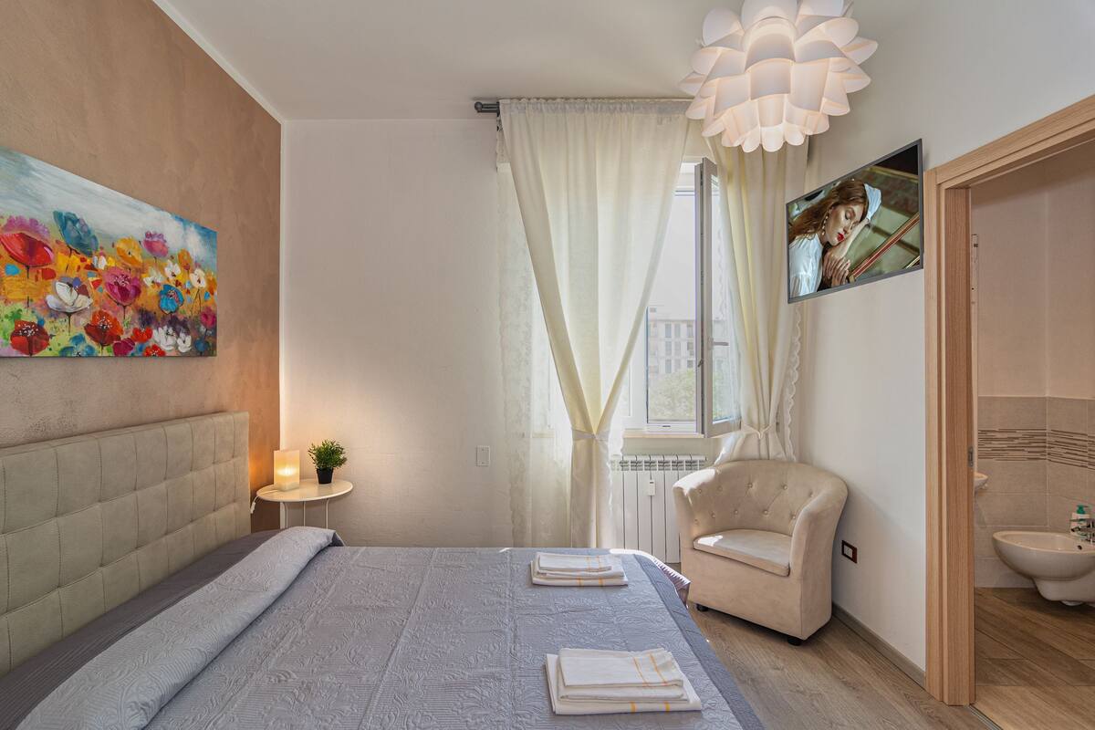 Un dormitorio con cama, una silla, un cuadro, una ventana con cortinas y un baño visible a través de una puerta abierta.