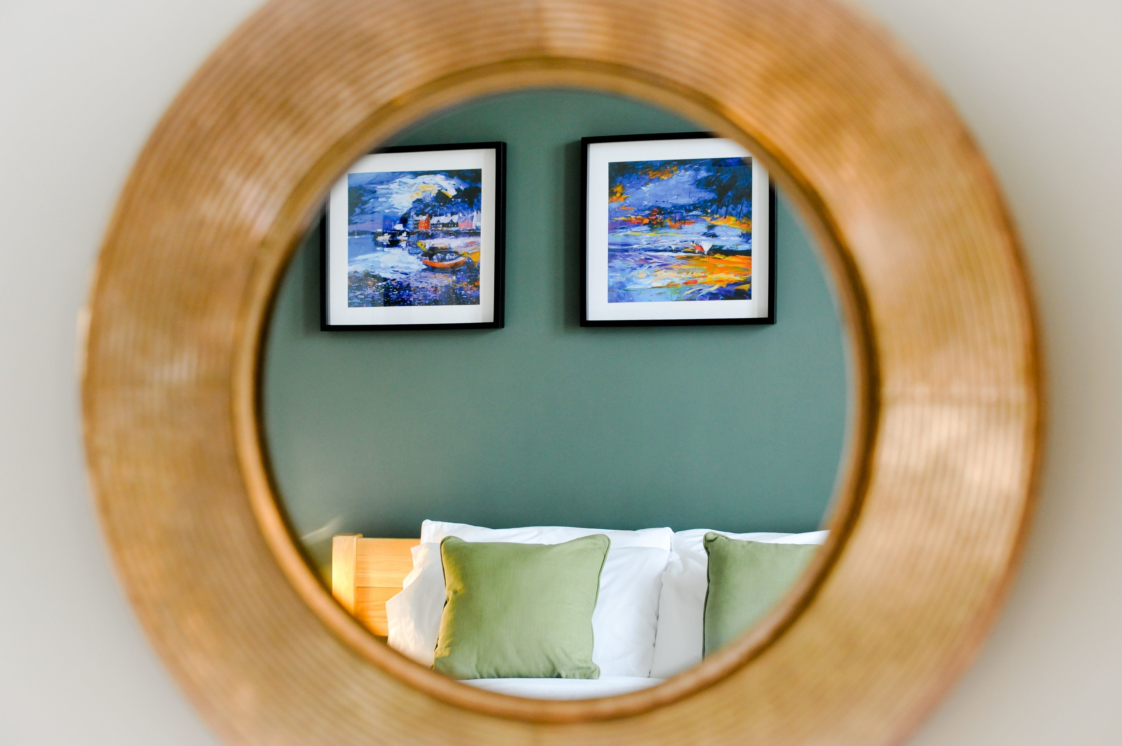 A circular mirror reflecting a bed with white and green pillows, and two framed paintings on the wall.