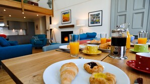 Ein Frühstückstisch mit Croissants, Saft und Kaffee auf einer Holztischplatte in einem modernen Wohnzimmer mit blauen Sofas und Kunstwerken an den Wänden.