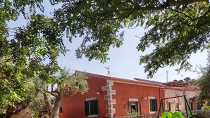 A playground with a slide and swings, surrounded by a stone wall and potted plants, in front of a red building with a stone facade. - Casale Imperatore (Ragusa)