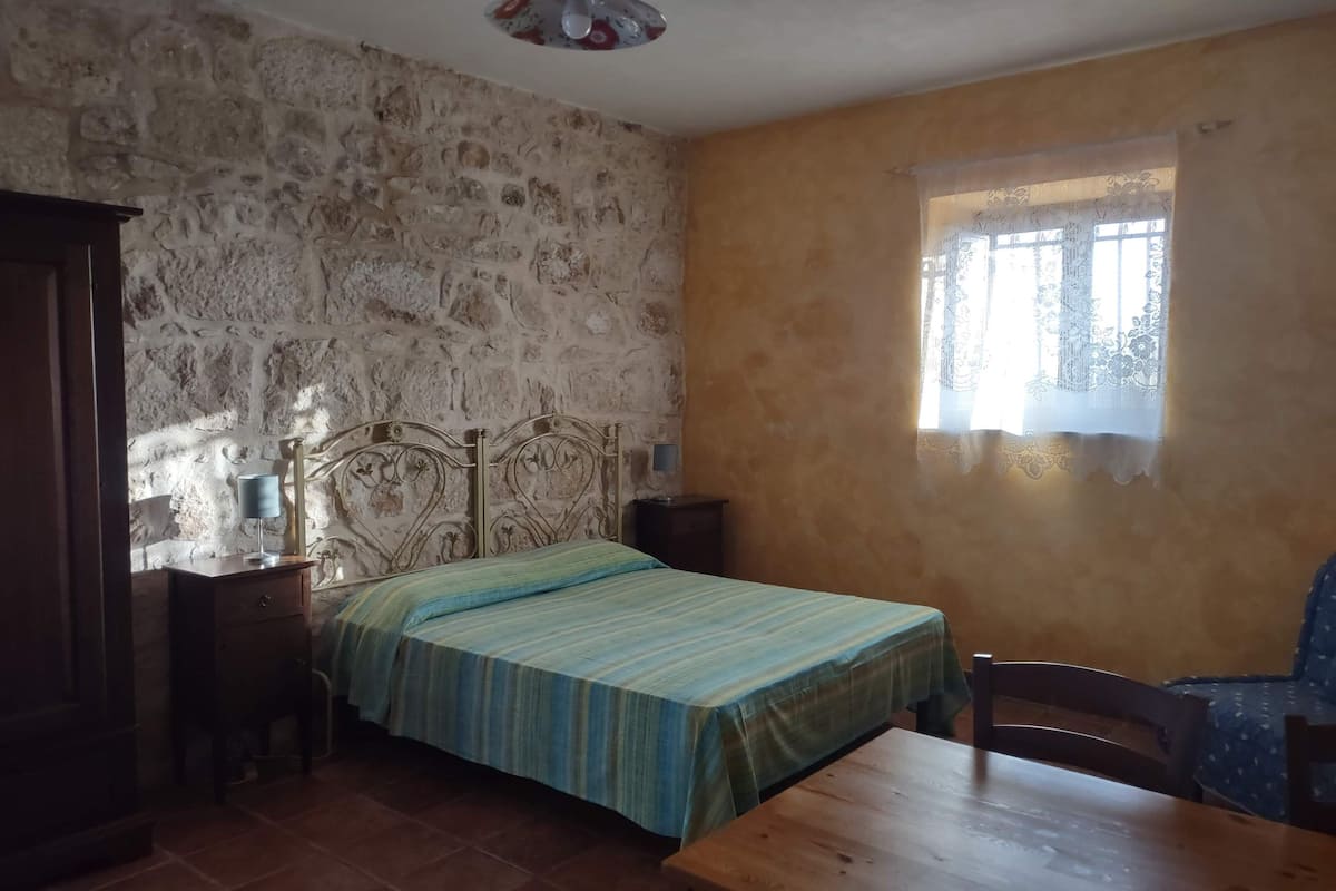 Ein Zimmer mit einem Bett, einem Holztisch, einem Stuhl, einem Nachttisch und einem Fenster mit Spitzenvorhängen.