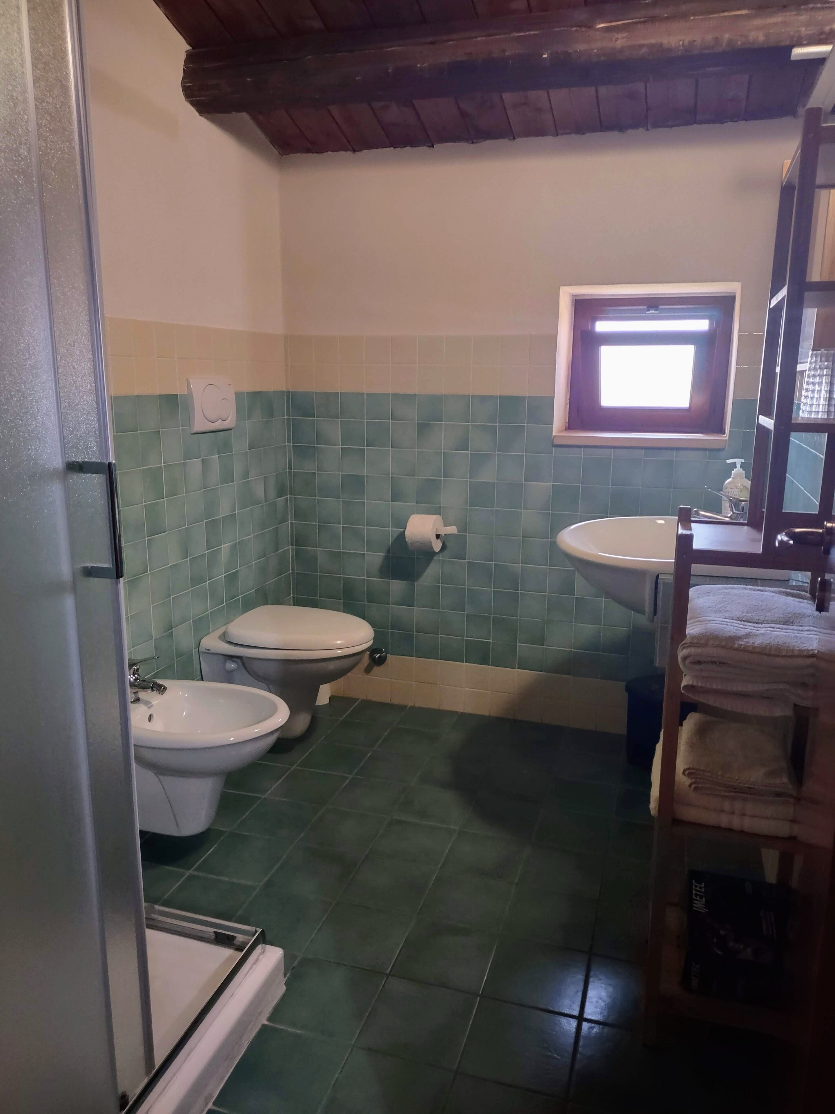 Casa de banho com bidé, sanita e lavatório, com azulejos em tons de verde e bege, e uma janela sobre o lavatório.