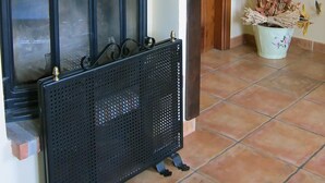 Une cheminée agrémentée d’un manteau décoratif, d’un paravent en métal noir et d’une plante en pot aux feuilles séchées.