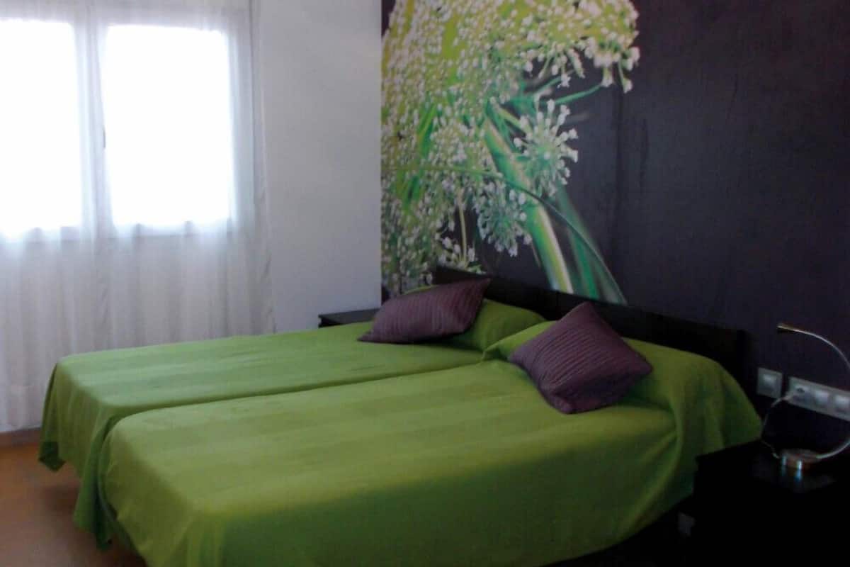 Une chambre avec deux lits recouverts de couvre-lits verts, un mur sombre orné d’une fresque florale et une fenêtre avec des rideaux blancs.