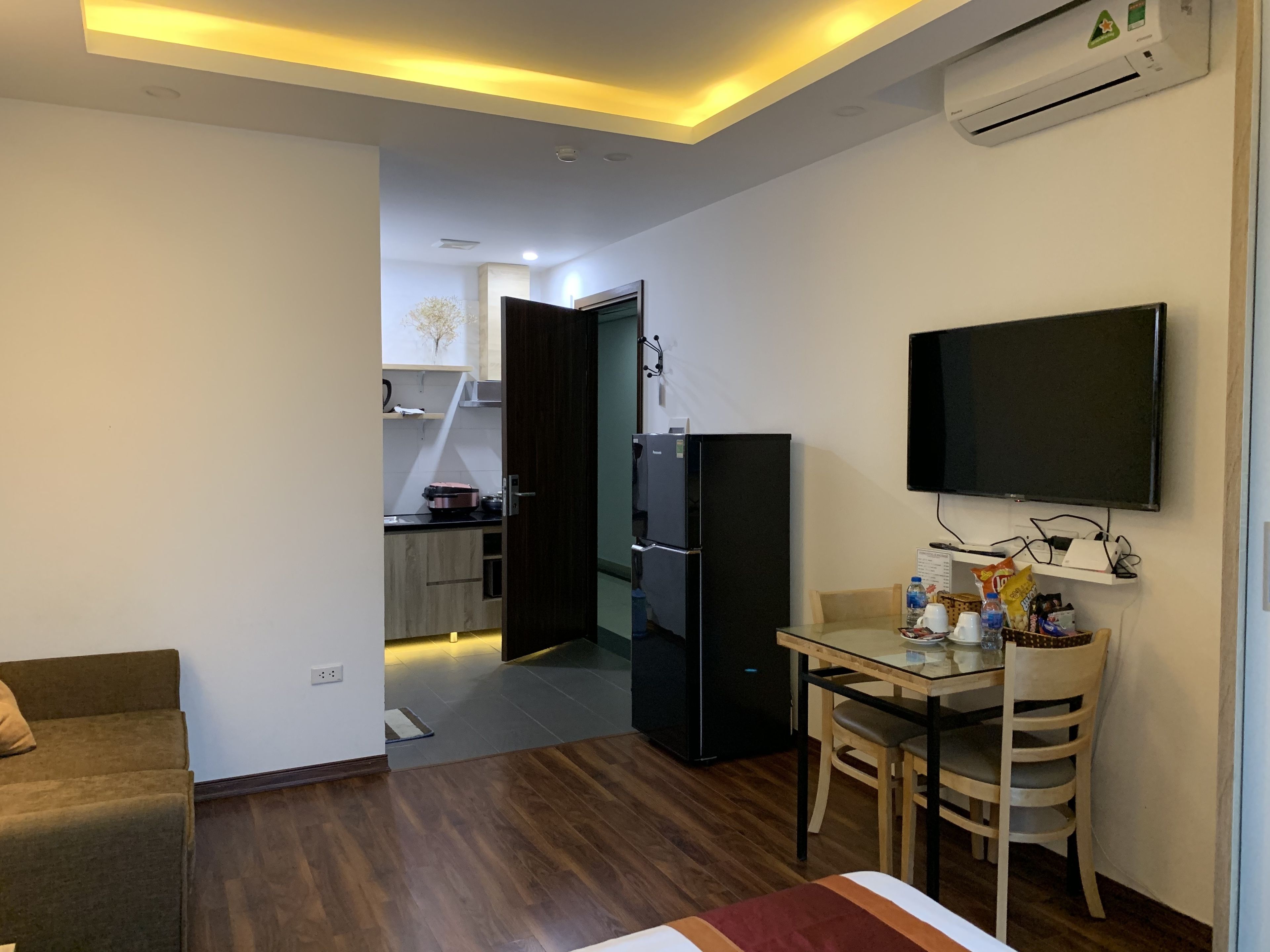 A modern living room with a sofa, a dining table with chairs, a flat-screen TV mounted on the wall, and a kitchen area visible through an open door.