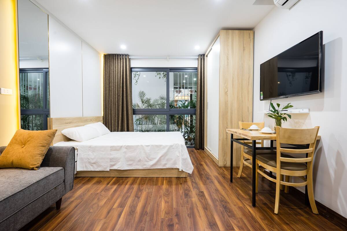 A modern hotel room with a bed, a sofa, a dining table with chairs, and a TV mounted on the wall.