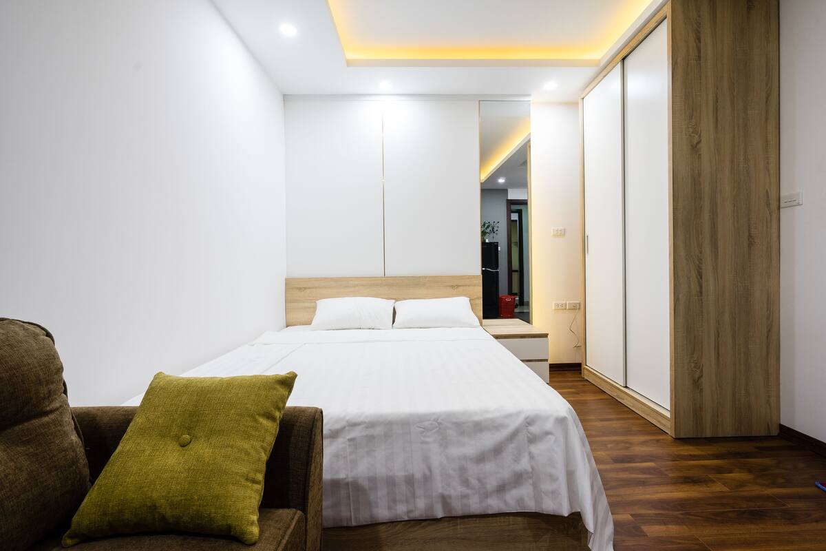 A modern hotel room with a neatly made bed, a wooden wardrobe, and a small seating area with a brown armchair and a green pillow.