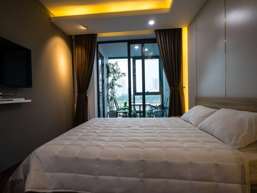 A hotel room with a large bed, a flat-screen TV mounted on the wall, and a view of the city through large windows.