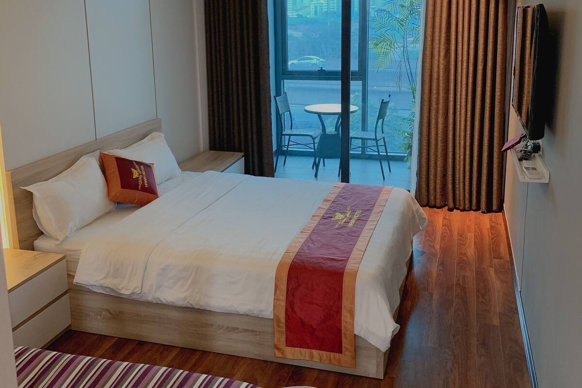 A hotel room with a large bed, a small table with chairs, a TV mounted on the wall, and a balcony with curtains.