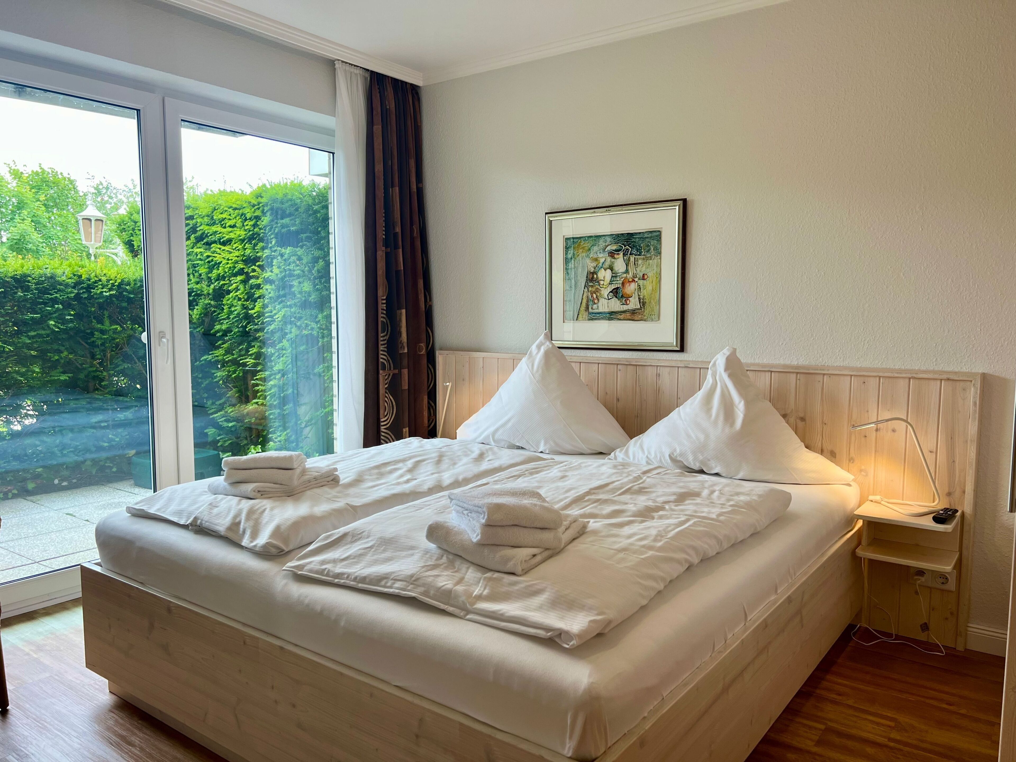 Un lit bien fait, avec du linge de lit et des serviettes blanches, une tête de lit en bois, une table de chevet avec une lampe et un tableau encadré au mur.