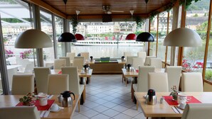 A restaurant with a view of a river and buildings across the water. Tables are set with white chairs, red placemats, and various dishes and drinks. - Hotel Haus Sonnschein (Cochem)