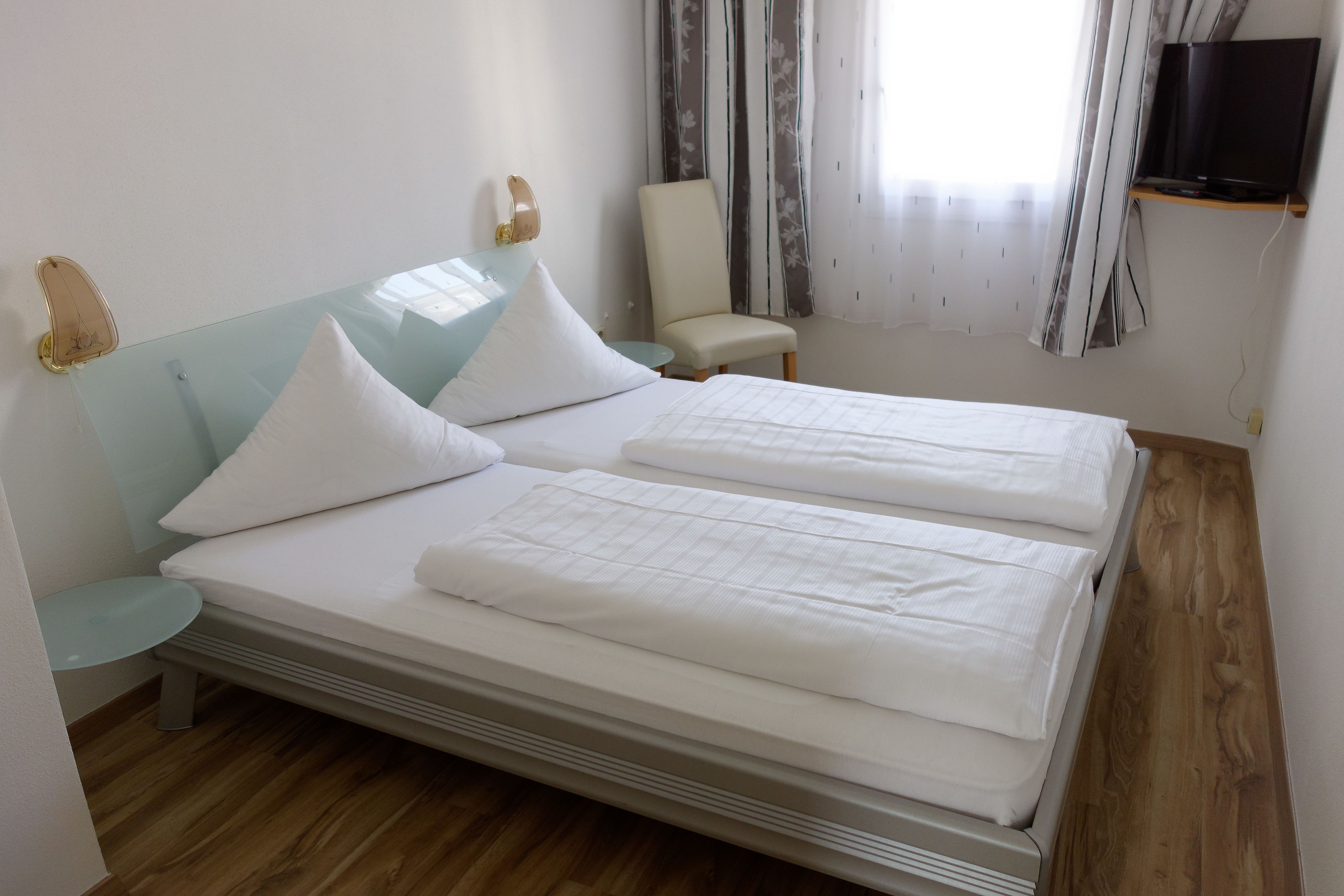 Una cama doble con ropa de cama blanca, una mesita de noche pequeña, una silla, un televisor sobre una estantería y una ventana con cortinas.