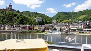 A riverside view of a town with historic buildings, a castle on a hill, and a table with a yellow tablecloth. - Hotel Haus Sonnschein (Cochem)