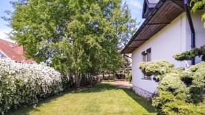 Un jardín bien cuidado con un árbol grande, arbustos recortados y un edificio blanco de techo inclinado.