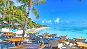 A beachside restaurant with wooden tables and wicker chairs, overlooking a clear blue ocean and a sandy beach lined with palm trees. - Seasense Boutique Hotel & Spa (Belle Mare)