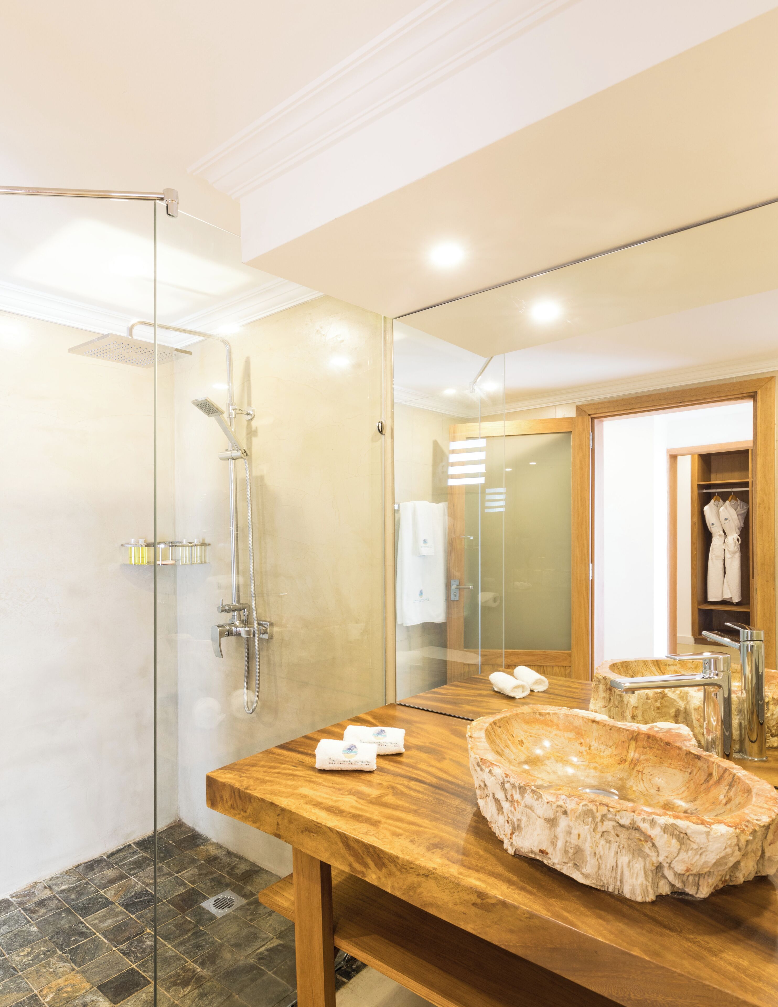 Un baño moderno con ducha de vidrio, tocador de madera y lavabo de mármol, y un espejo grande que refleja el ambiente.
