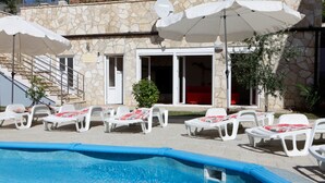 Un espace piscine avec des chaises longues et des parasols, un bâtiment en pierre doté d’une porte et de fenêtres, et un escalier menant à un niveau supérieur.