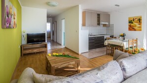 Un salon moderne avec un téléviseur à écran plat, une table basse en bois et un canapé. On aperçoit à l’arrière-plan un coin repas avec une table et des chaises.