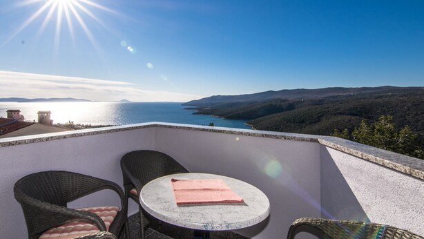 Un balcon agrémenté de fauteuils en osier et d’une petite table ronde, offrant une vue panoramique sur un plan d’eau et des collines.