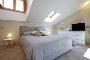 Un dormitorio con cama, un escritorio con televisor, una mesita con lámpara y un tragaluz.