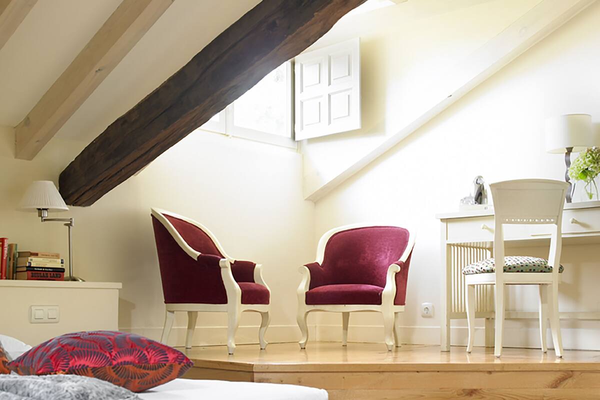 A bedroom with a sloped ceiling, wooden beams, a bed with patterned bedding, two red armchairs, a white desk with a chair, and a radiator.