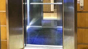 Un ascensor con paredes metálicas y una puerta de vidrio, que da a un pasillo con una puerta de madera al final.