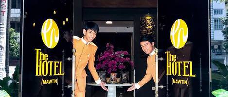 Entrada de hotel con puertas de cristal, mostrador de recepción y una planta en maceta.
