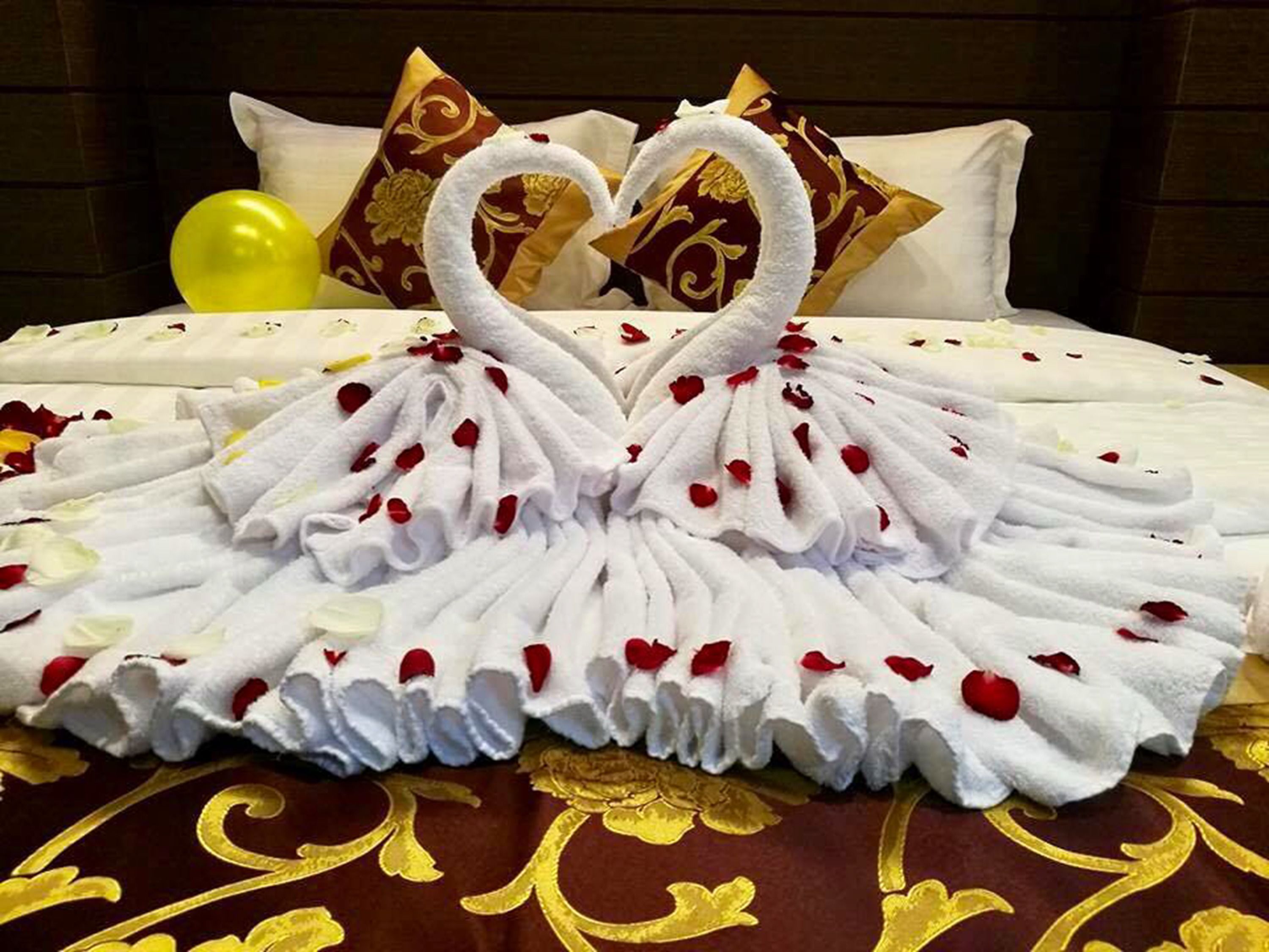 A bed with a heart-shaped towel arrangement and rose petals.