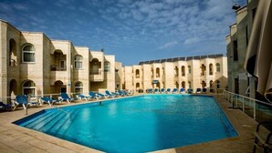 Ein Hallenbad mit blauem Wasser, umgeben von beige gefärbten Gebäuden mit Bogenfenstern und Balkonen.