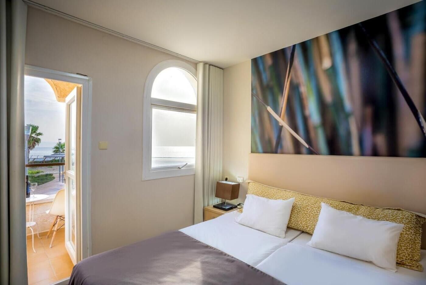 A hotel room with a bed, a nightstand, a lamp, a window with a view of the beach, and a painting on the wall.